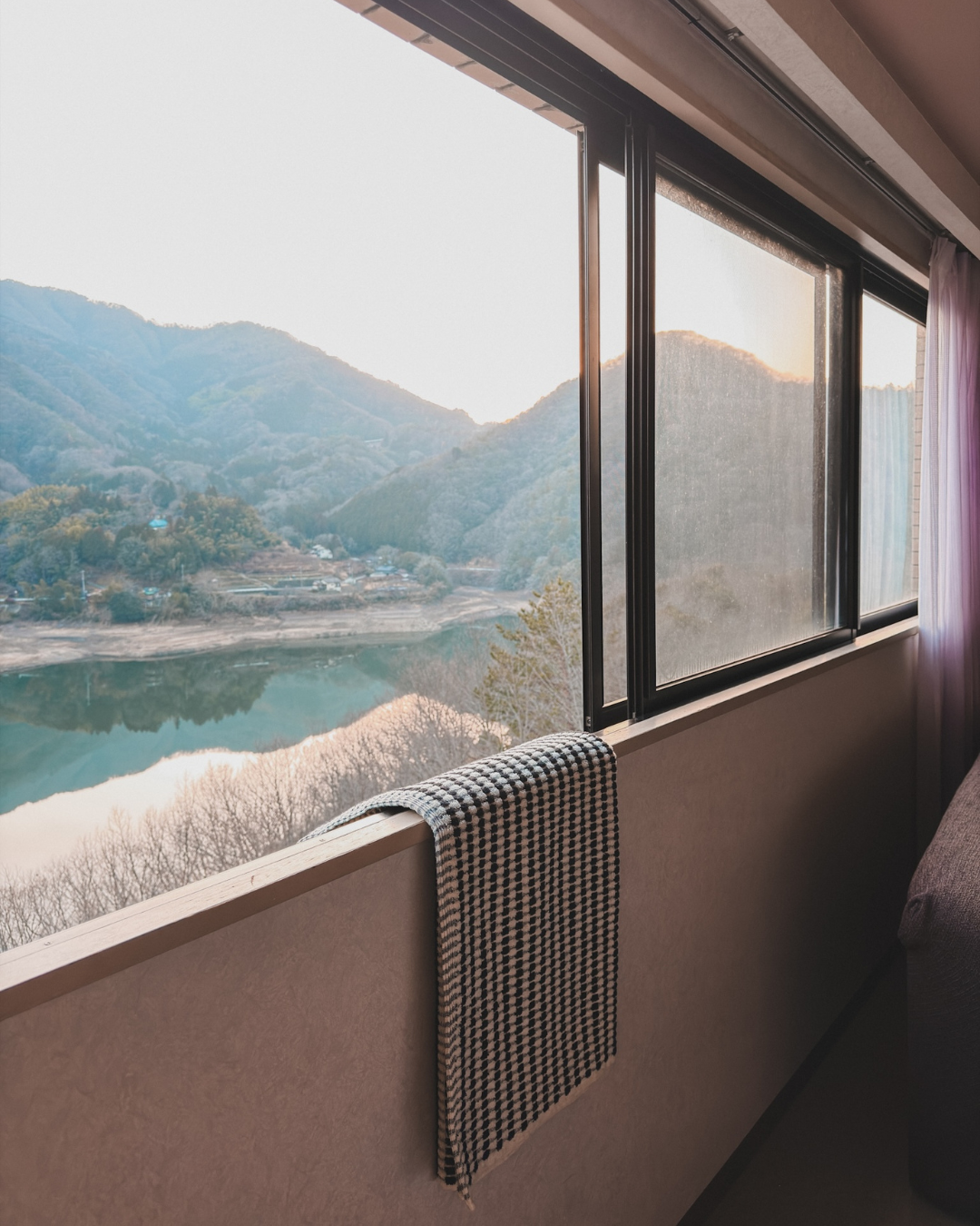 A room with a large window offering a view of mountains and a river while monochrome textured bath towel by mandisgn